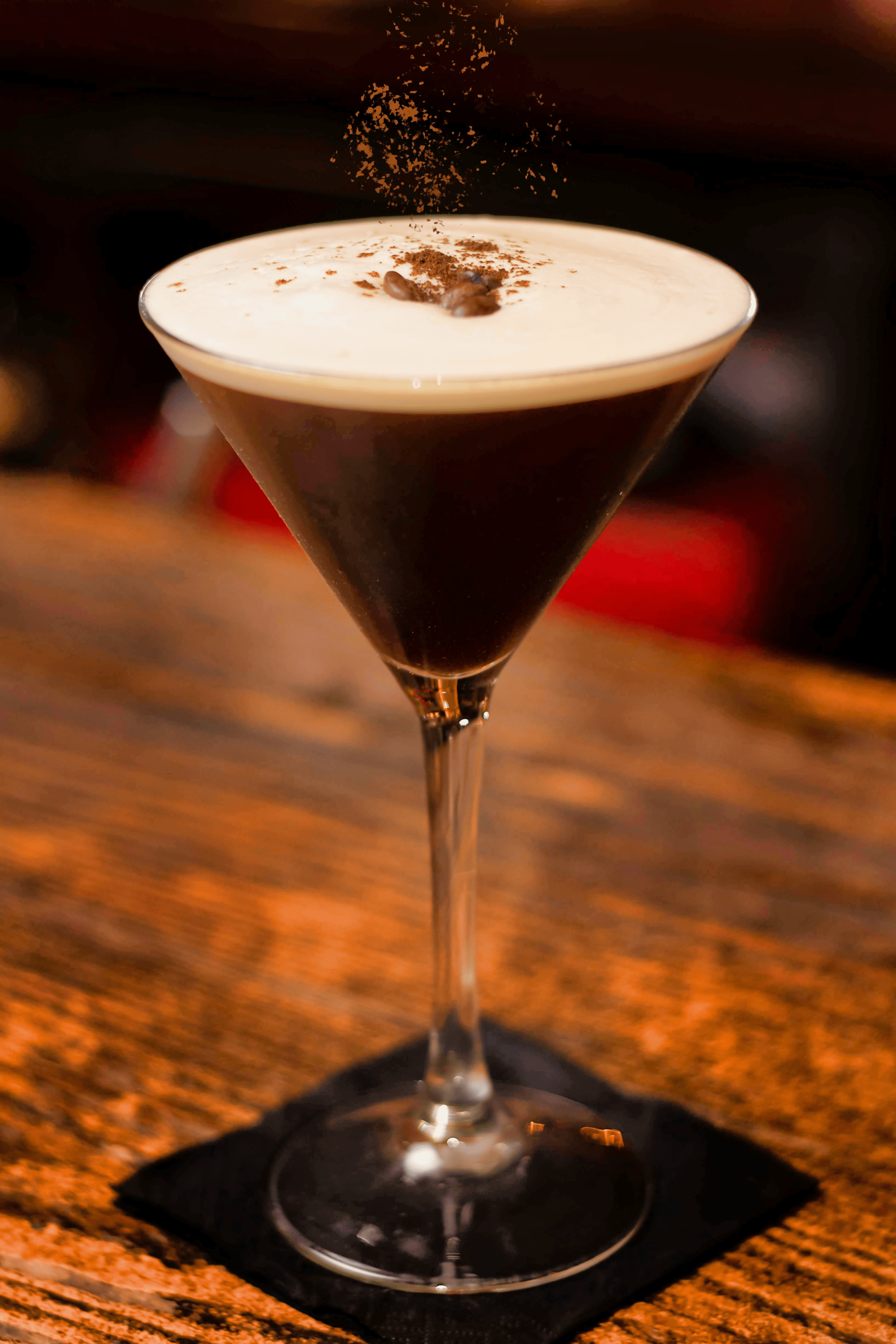 An Espresso Martini glass topped with coffee beans being garnished with chocolate shavings raining down on the creamy surface.