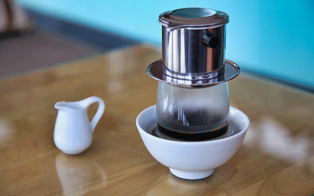 A Vietnamese coffee being brewed with a phin over a clear glass which is placed in a small bowl filled with water to quickly cold down the coffee.